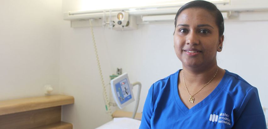 Beena Sasi is standing in a patient room in front of a bed and some medical equipment. She is smiling and is wearing a blue Mater Private Network uniform.