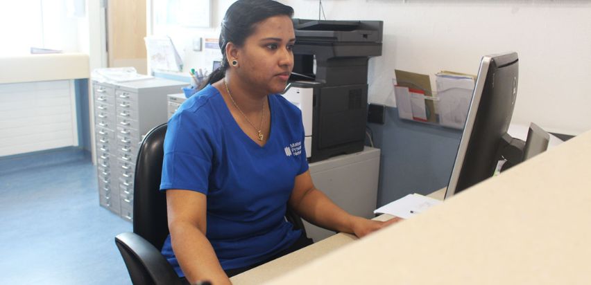 Beena Sasi is sitting at a nurses station and is using a computer which is positioned in front of her.