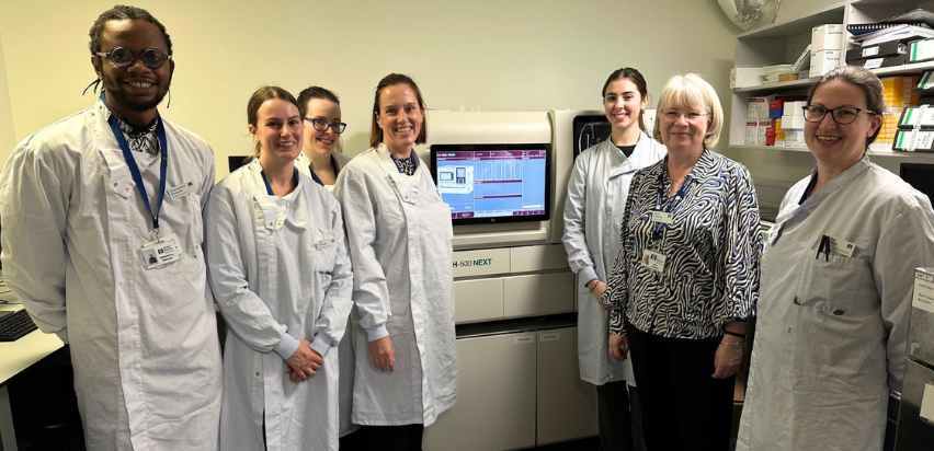Medical Scientists are standing together for a group picture. They are all wearing long white lab coats and are standing beside a machine that is used to analyse patient samples.