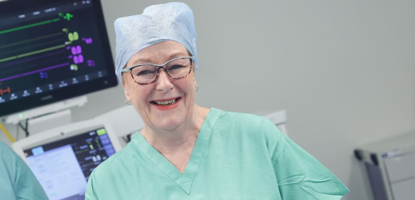 Deirdre Crosskill is smiling while standing in front of medical equipment in an operating theatre. She is wearing glasses, a green top and a blue theatre cap.