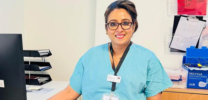 Jincy Kuriakose is smiling and and sitting in front of a computer screen. She is wearing glasses, a light green top and has a lanyard around her neck.