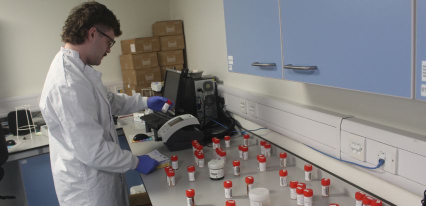 A male medical scientist is standing at a counter top in the laboratory. He is looking at a sample in a small container with a red lid and comparing the information on the container with information on a sheet of paper.