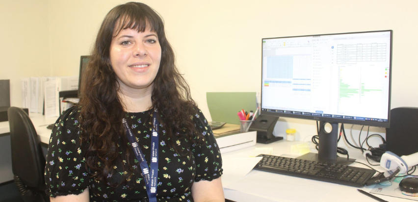 Maite Devos Lozano is sitting at a desk in front of a computer screen and keyboard. She is wearing a black dress with a flowery pattern and has long brown hair.