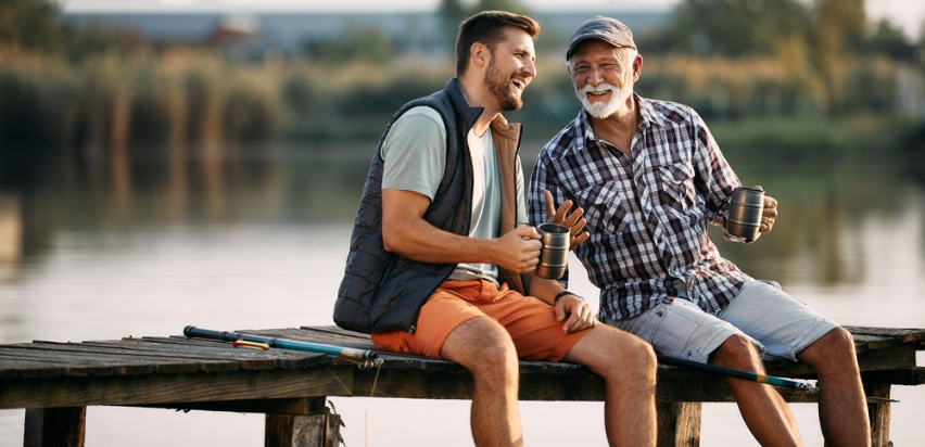 A younger man and an older man are sitting together at the end of a jetty on a lake. They are leaning toward each other and are lauging. They have fishing rods beside them and are holding a cup each.