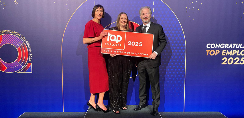 Two women and one man are standing on a stage holding a red sign which has Top Employer branding on it in white letters.