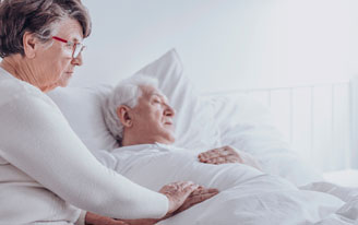 visit-a-patient A woman sitting at the bedside of her husband who is asleep. She has her hand on his hand.