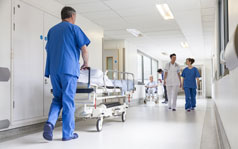 your-hospital-stay Various medical professionals walking down the corridor of a hospital. One man is wheeling a patient bed.