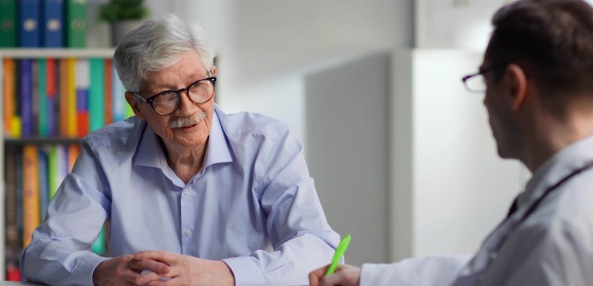 An older man sitting at a desk speaking to a doctor